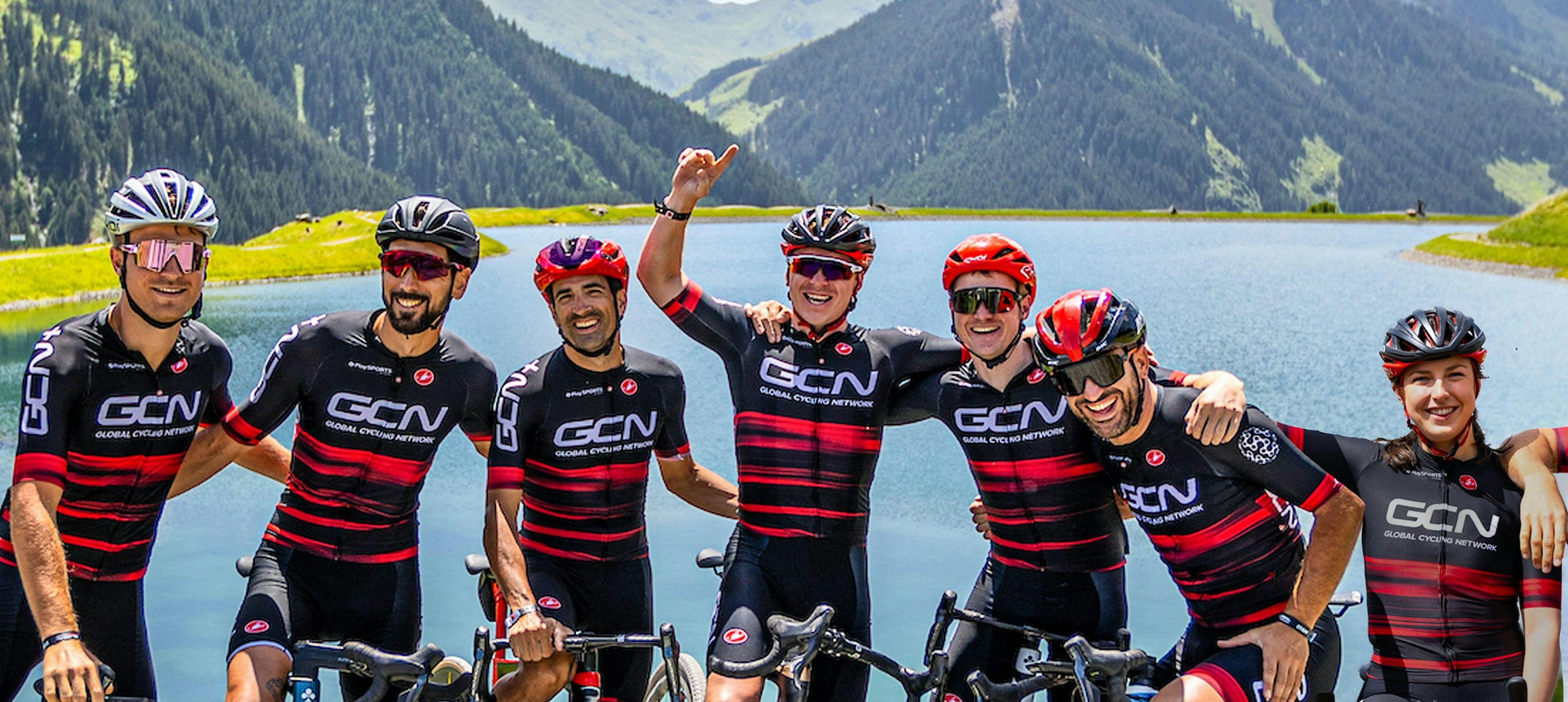 7 GCN presenters in GCN-branded cycling kit smile and laugh in a group whilst holding bikes. There's mountains and a lake in the background